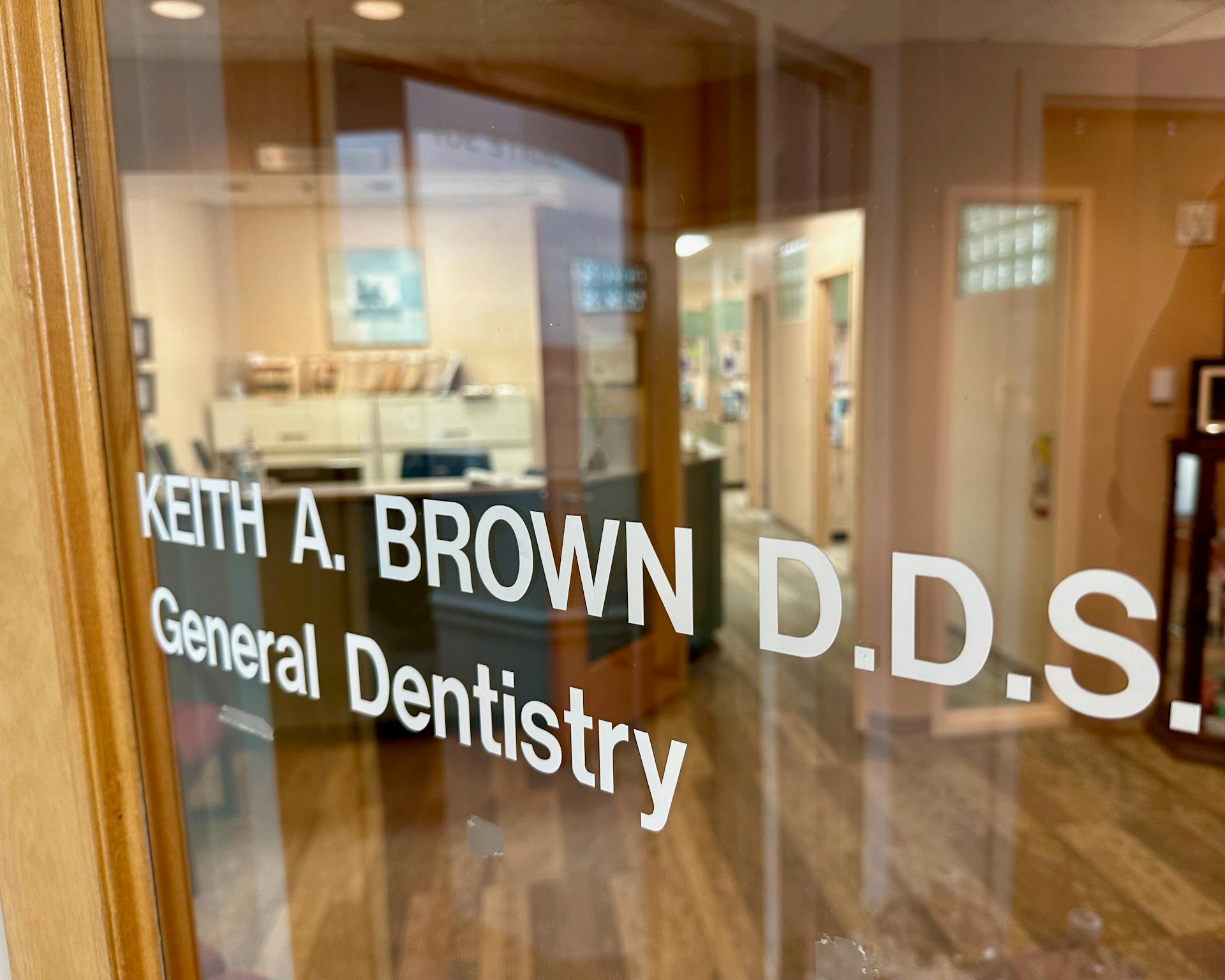 Dr. Keith A. Brown DDS, FAGD, standing with arms crossed in his office at 1295 Rickert Drive, 3rd floor, Naperville, IL, while a staff member is seated at a desk in the background.