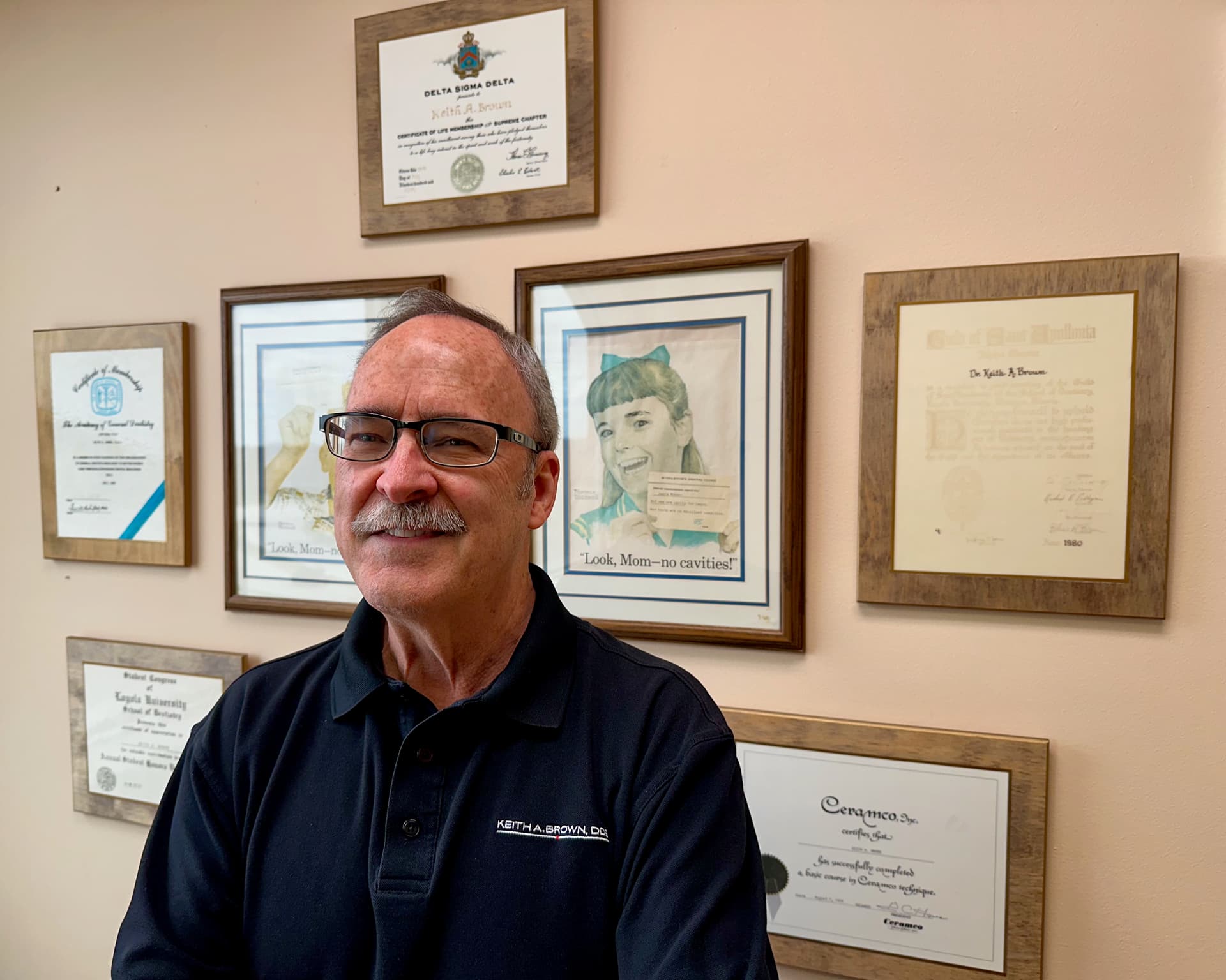 Dr. Keith A. Brown DDS, FAGD, standing in front of framed certificates with the Fifth Third Bank building at 1295 Rickert Drive, 3rd floor, Naperville, IL, in the background.