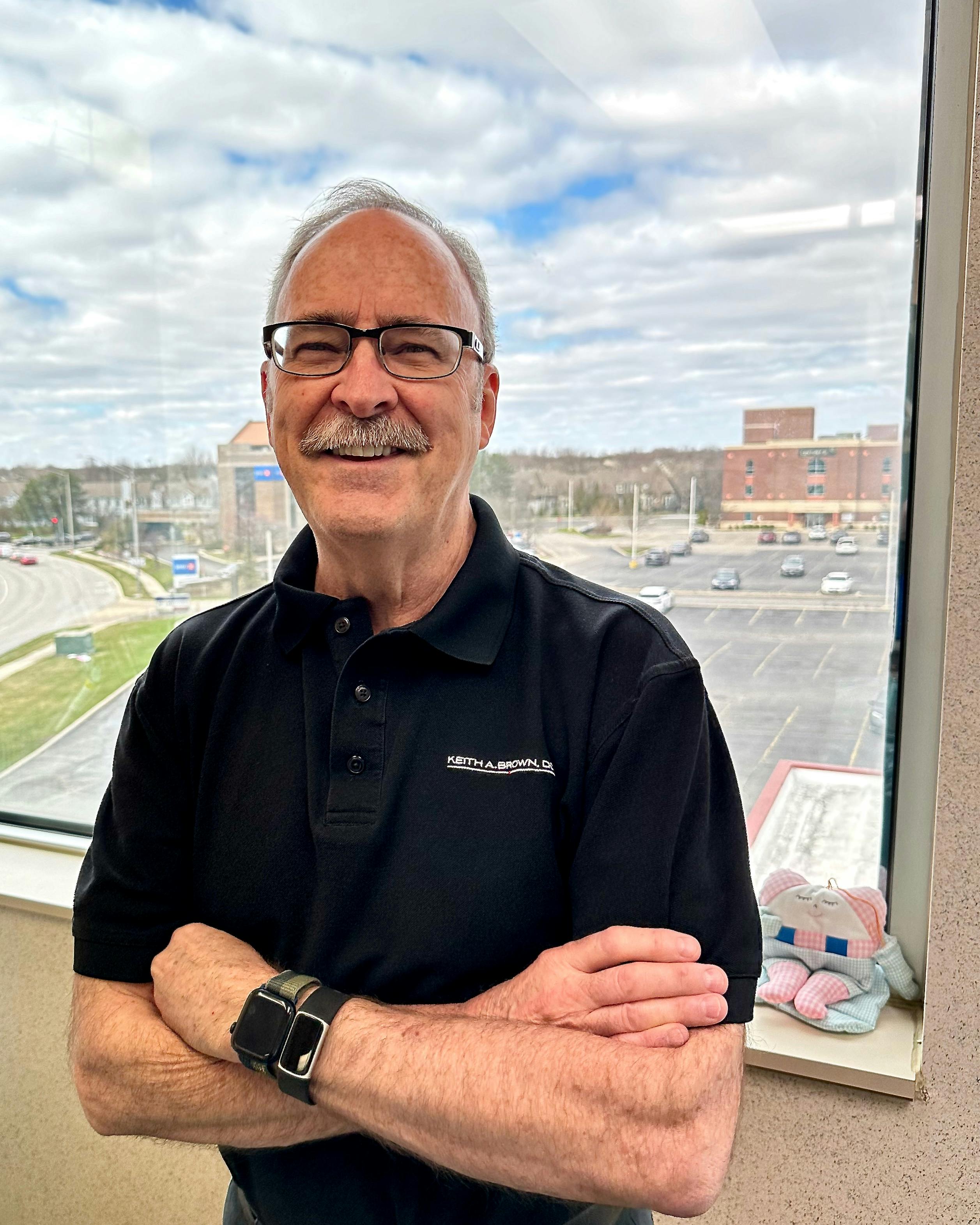 Portrait of Dr. Keith A. Brown DDS, FAGD, with a view of the Fifth Third Bank building and surrounding area in Naperville, IL, from the third floor.