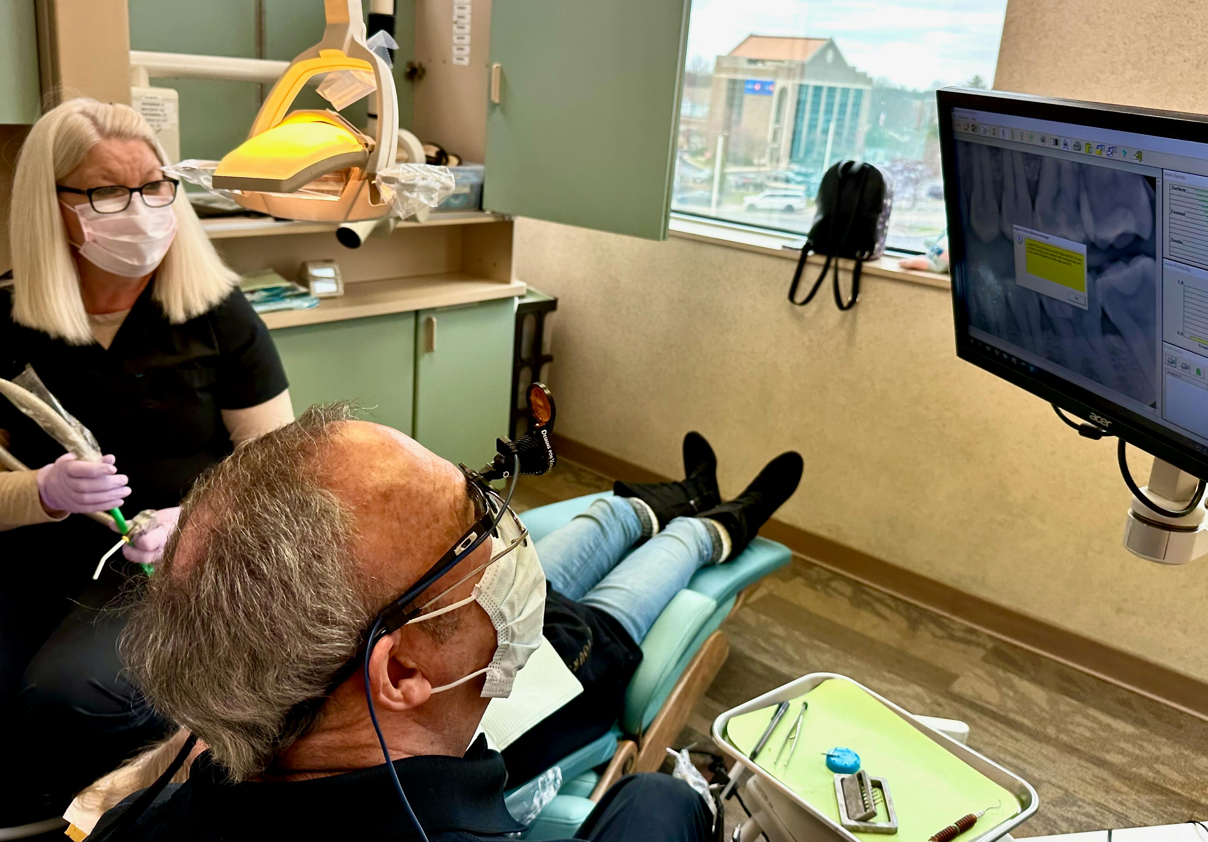 Dr. Keith A. Brown DDS, FAGD, with a patient and a computer displaying dental images in the background.